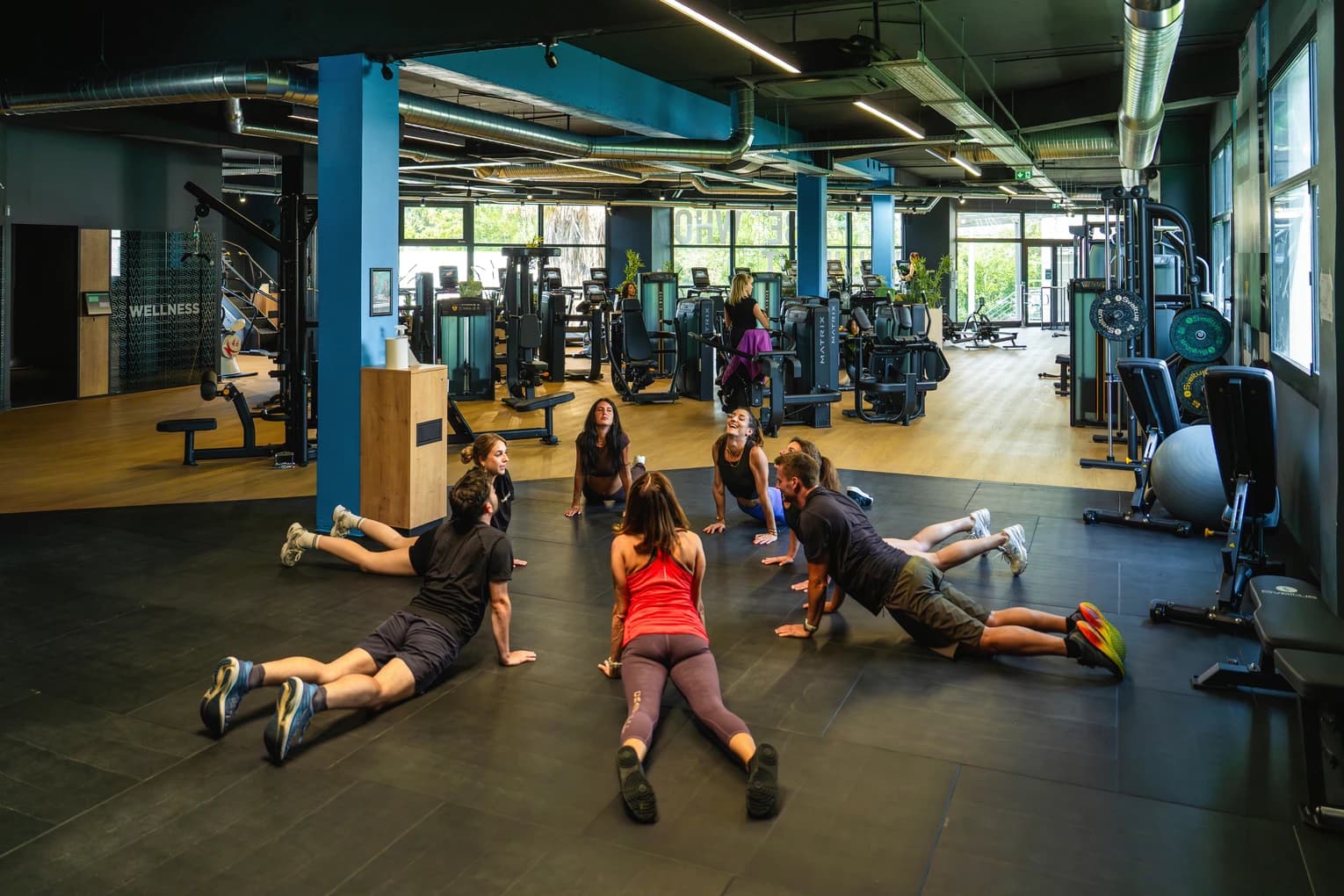 Salle de sport Keepcool, groupe en cours collectif mobilité au sol avec exercices variés, équipements cardio et musculation en arrière-plan.